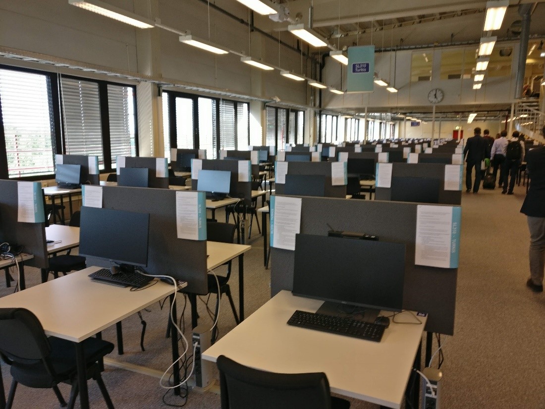 Photograph of NTNU Eksamen (exam) building interior set up for exams with screened desks with computers and instructions pinned to the desk screens in long rows